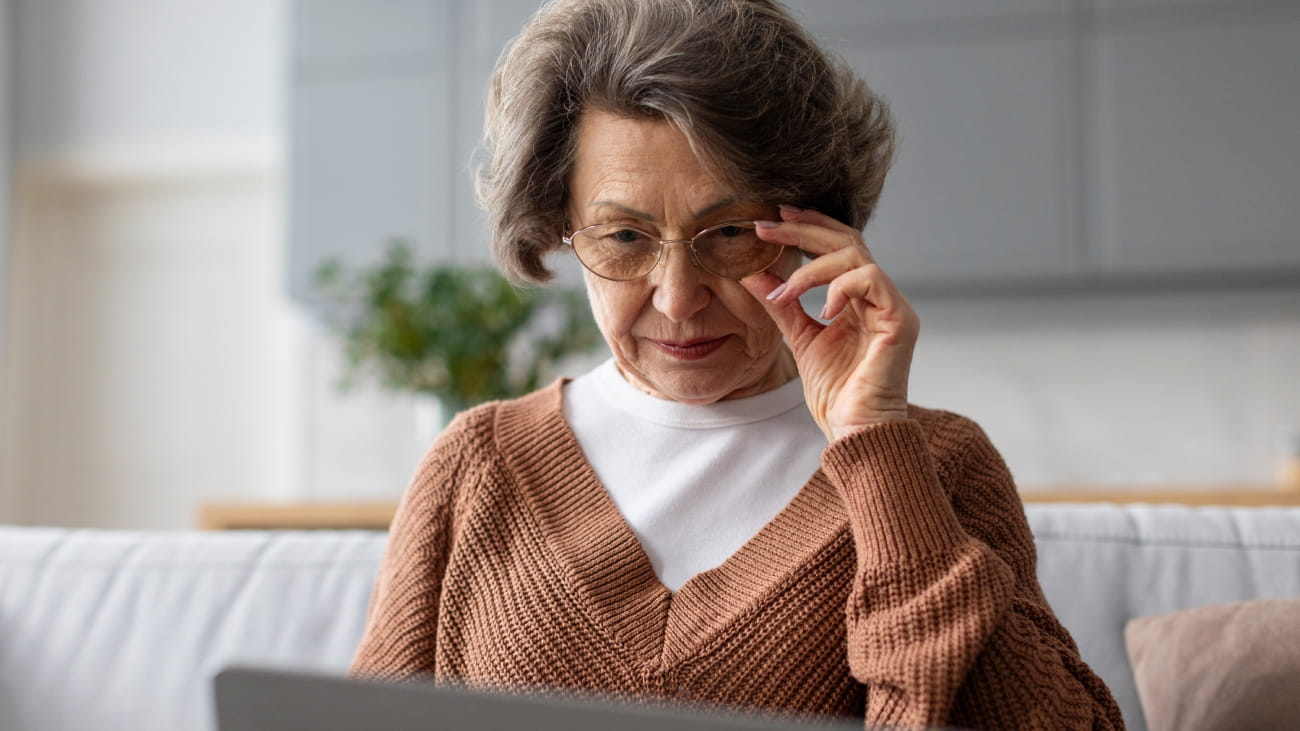 Senior woman using laptop and touching glasses, surfing internet, having problems with vision, sitting on sofa at home interior.