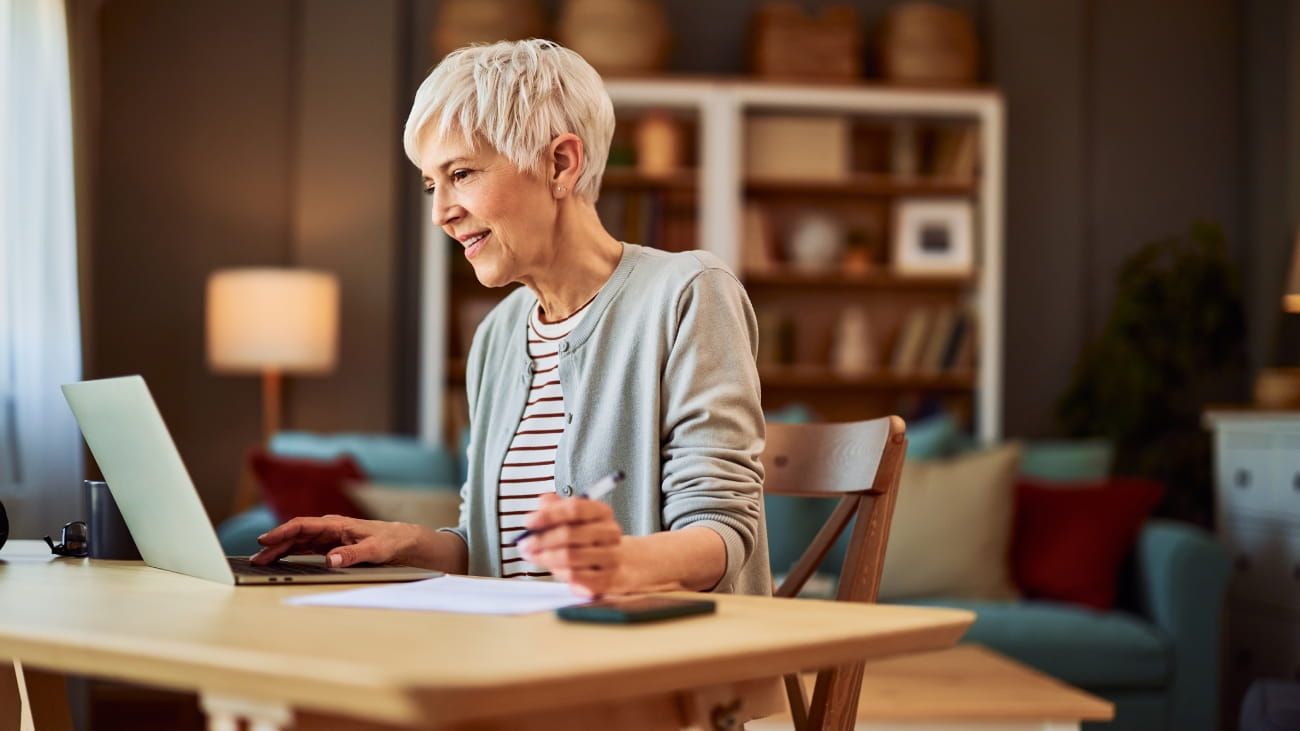 A professional and focused senior adult female freelancer working from home using a laptop and taking notes on a paper.