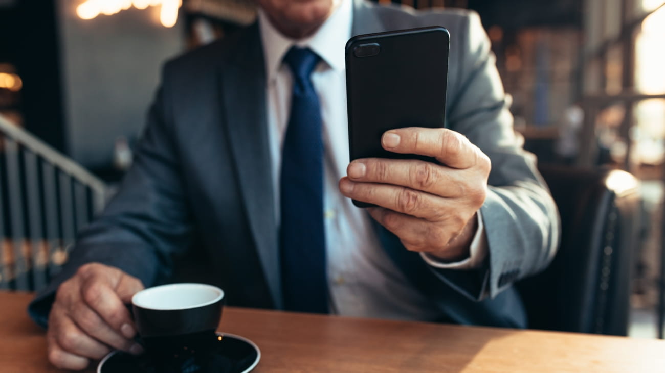 Close up of mobile phone in hand of a businessman with coffee on cafe table. Senior businessman using smartphone at coffee shop.