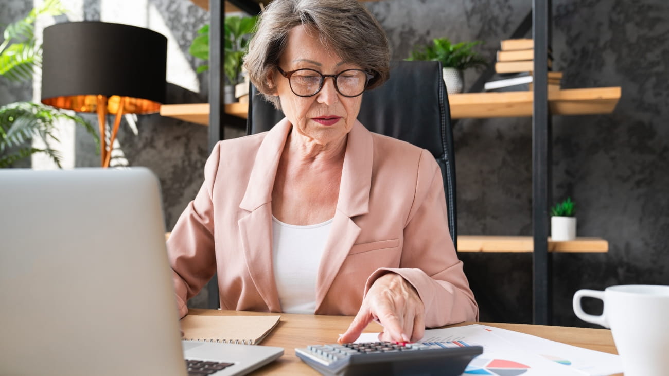 Concentrated caucasian senior businesswoman manager counting funds earnings bills on calculator in office. Serious grandmother paying domestic bills, loan, mortgage, insurance.