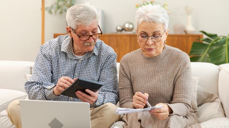 Middle aged senior couple sit with laptop and paper document. Older mature man woman reading paper bill pay online at home managing bank finances calculating taxes planning loan debt pension payment.