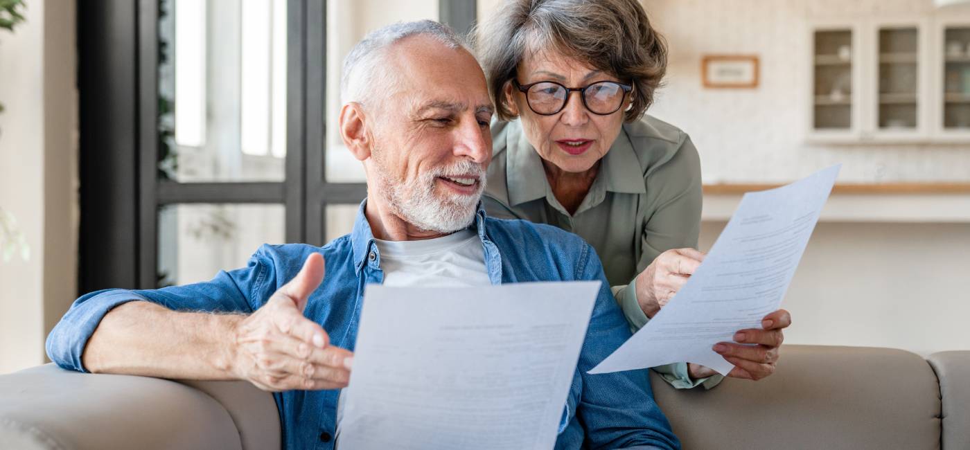 A senior couple, man sits on a sofa, his wife leaning over, reviewing taxes together, focused and engaged in their living room