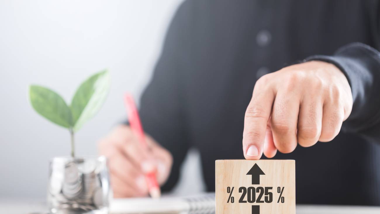 A businessman points at wooden blocks while a coin jar with a sprouting plant sits nearby. He notes numbers in a notebook, symbolising analysis of 2025 interest rate trends-rise or fall