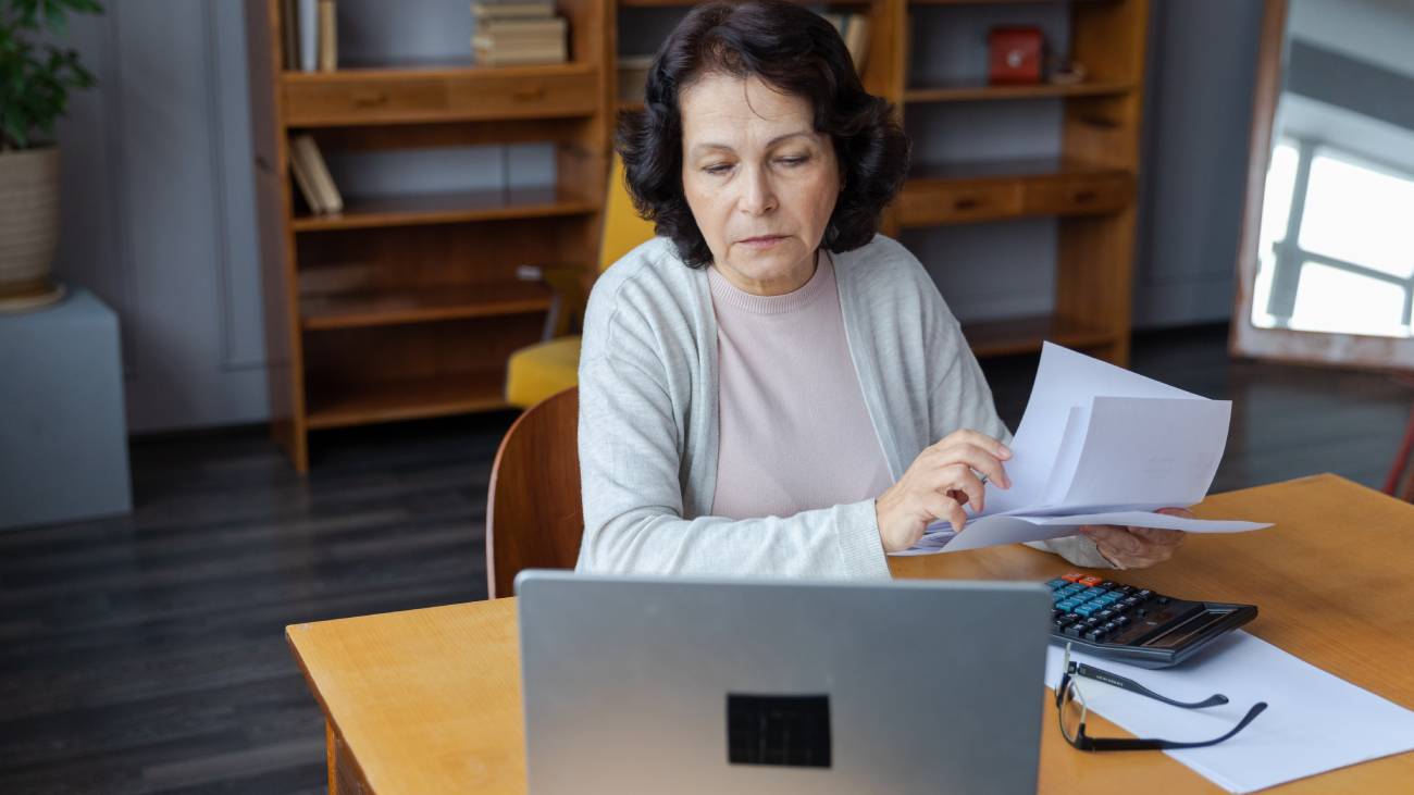 Middle aged senior woman sit with laptop and paper document,  reviewing pension payments