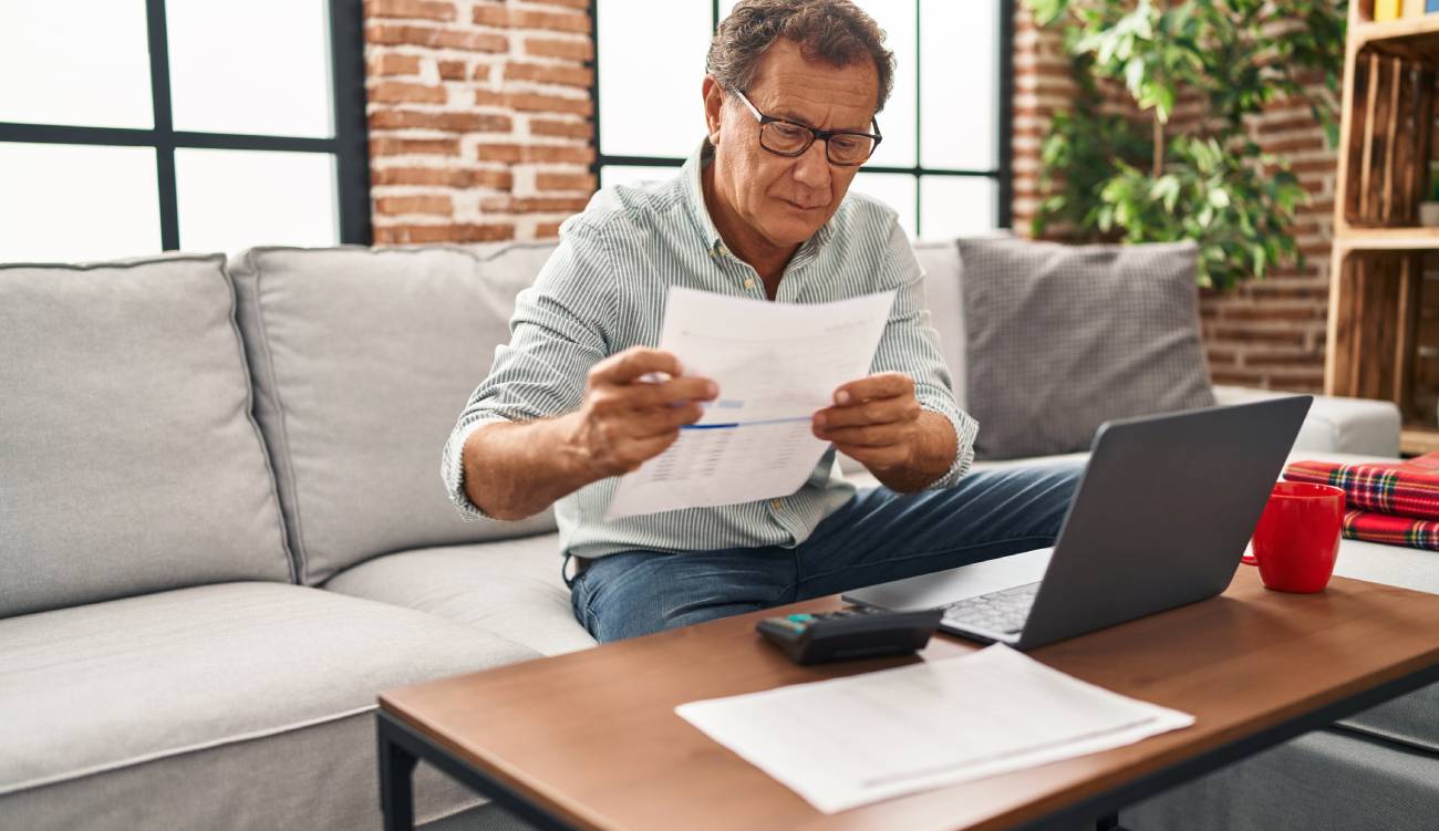 A mature man sitting on a sofa, reading a letter with his laptop open and calculator on a table.
