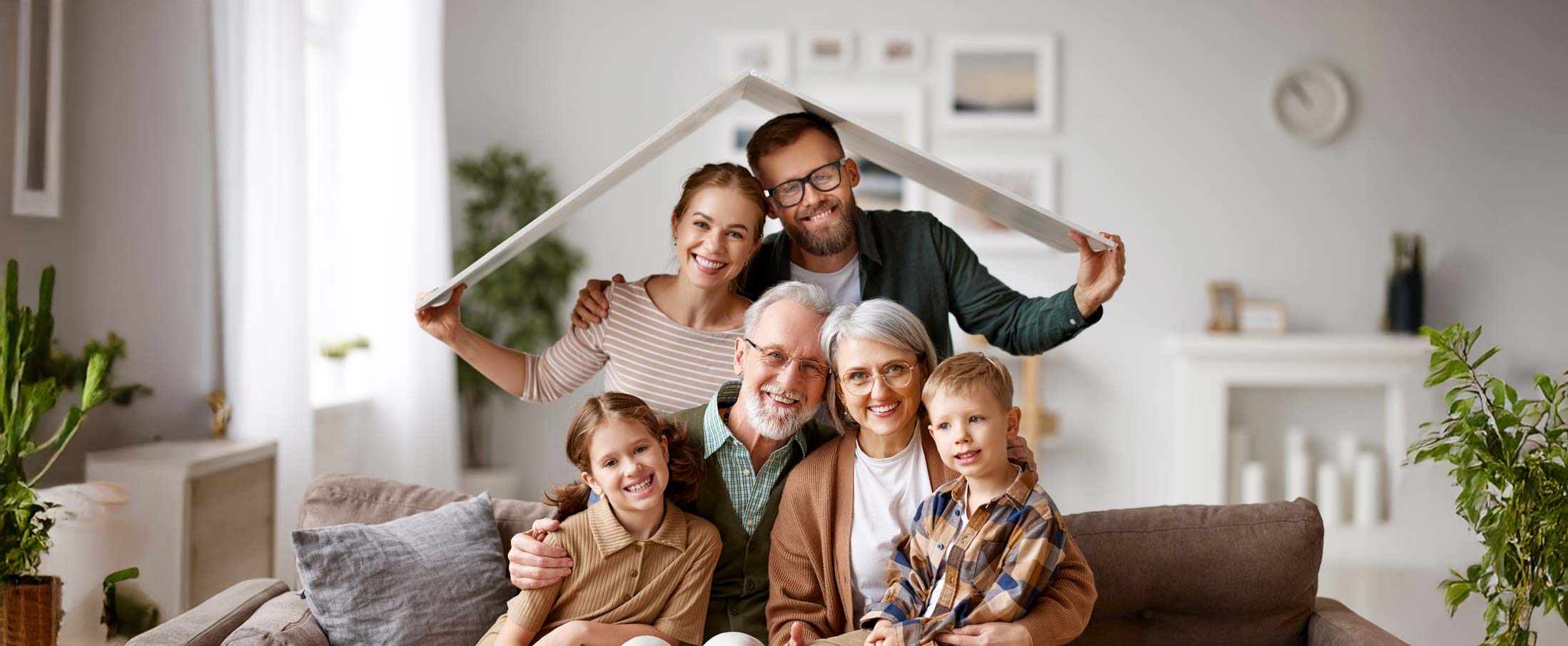Big happy family, grandparents, mother, father with little kids son and daughter celebrating relocation in new home, sitting on the coach under paper roof and smiling at camera. Mortgage loan concept