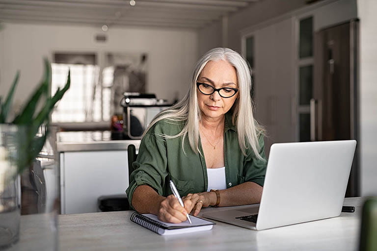 Senior woman taking notes in notebook while using her laptop at home