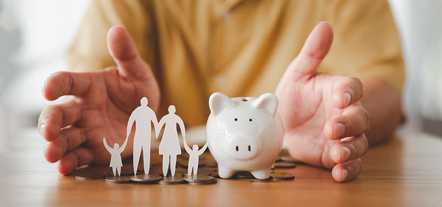 A pair of hands open on a table with a paper cutout family and a piggy bank between them
